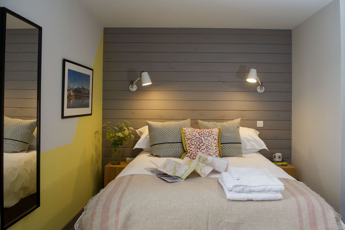 Una cama bien hecha con almohadas, una mesita de noche con lámpara, un cuadro en la pared y un espejo.