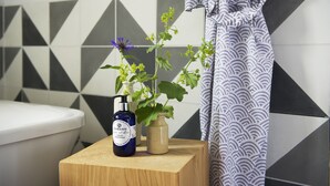 A bathroom with a toilet, a wooden stool, a bottle of hand soap, a vase with a plant, and a patterned shower curtain.