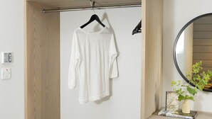 A room with a wooden wardrobe, a hanging rack with clothes, a black chair, and a small table with a plant and a book.