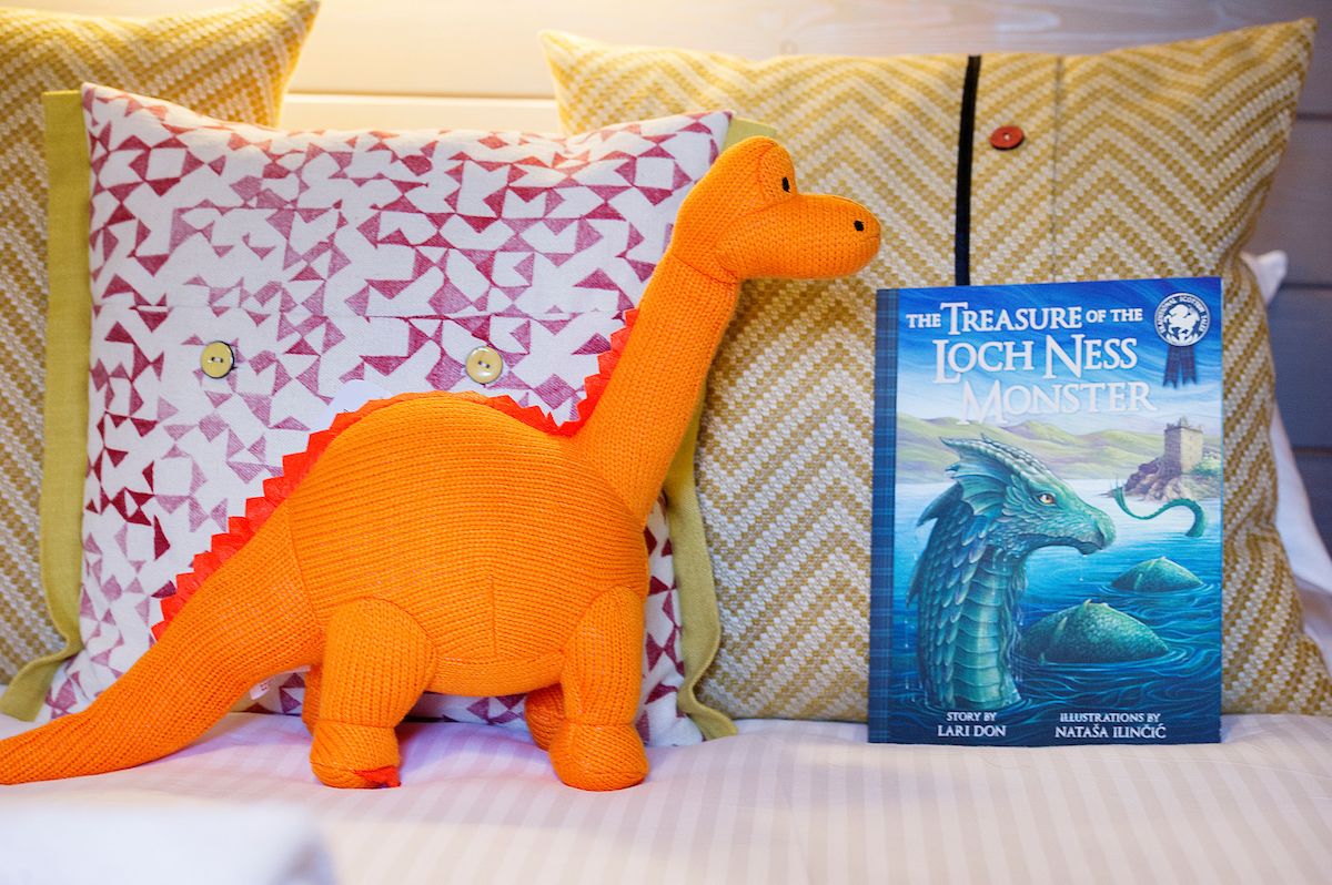 A plush dinosaur toy and a book titled "The Treasure of the Loch Ness Monster" on a bed with patterned pillows.