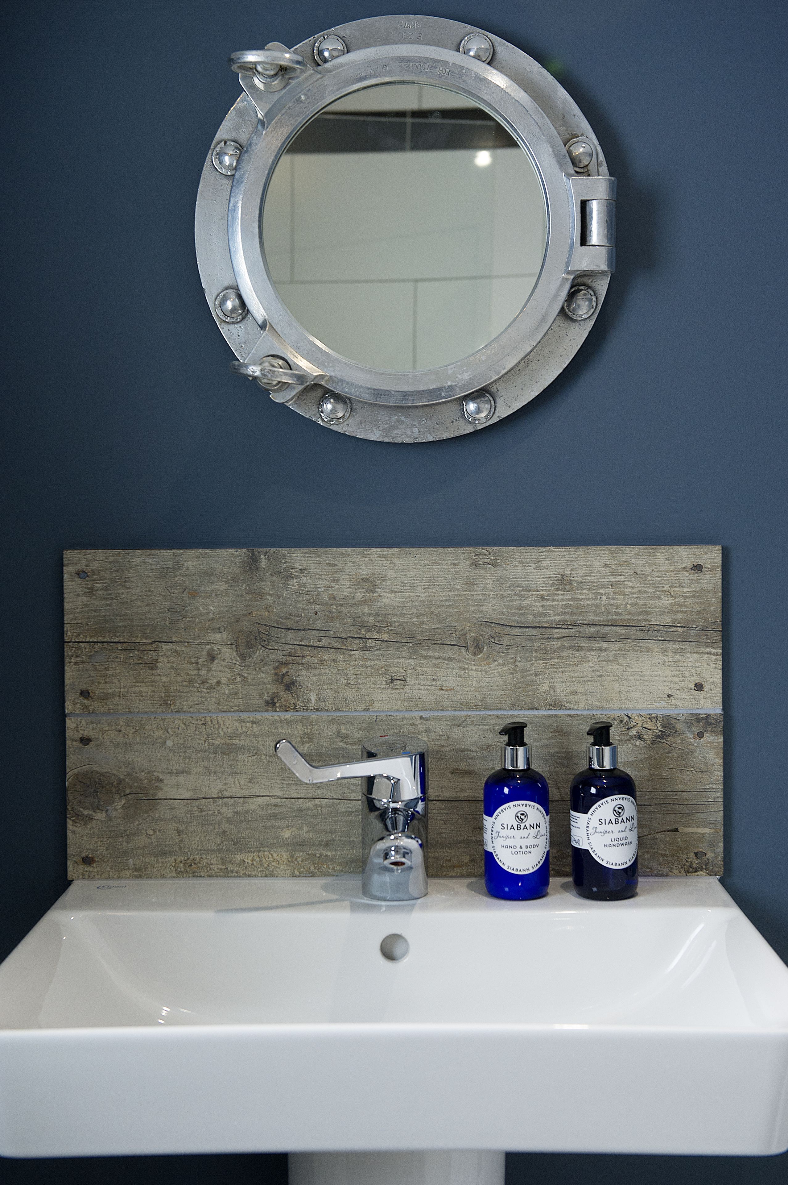 A bathroom with a round mirror, a wooden panel above the sink, and two hand and body lotion bottles.