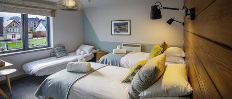 A hotel room with two beds, a window with a view of houses, a small table, and a lamp.