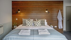 A neatly made bed with a striped blanket, two pillows with a geometric pattern, and two folded towels on the bed. There are two wall-mounted lights on either side of the bed, a small table with a vase and a cup, and a towel hanging on the wall.