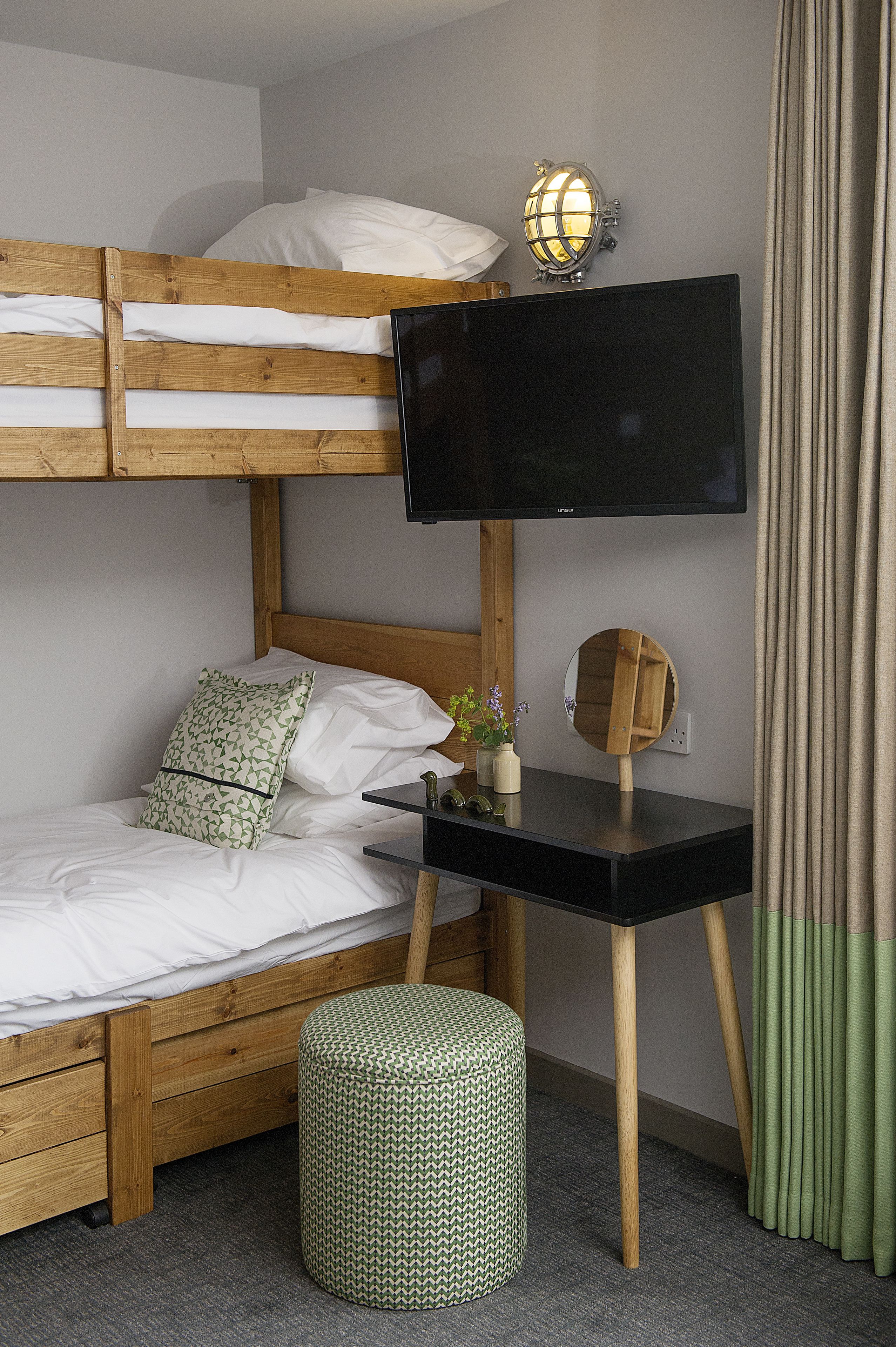 Una cama litera con un televisor encima, una mesita con un espejo y un otoman verde.