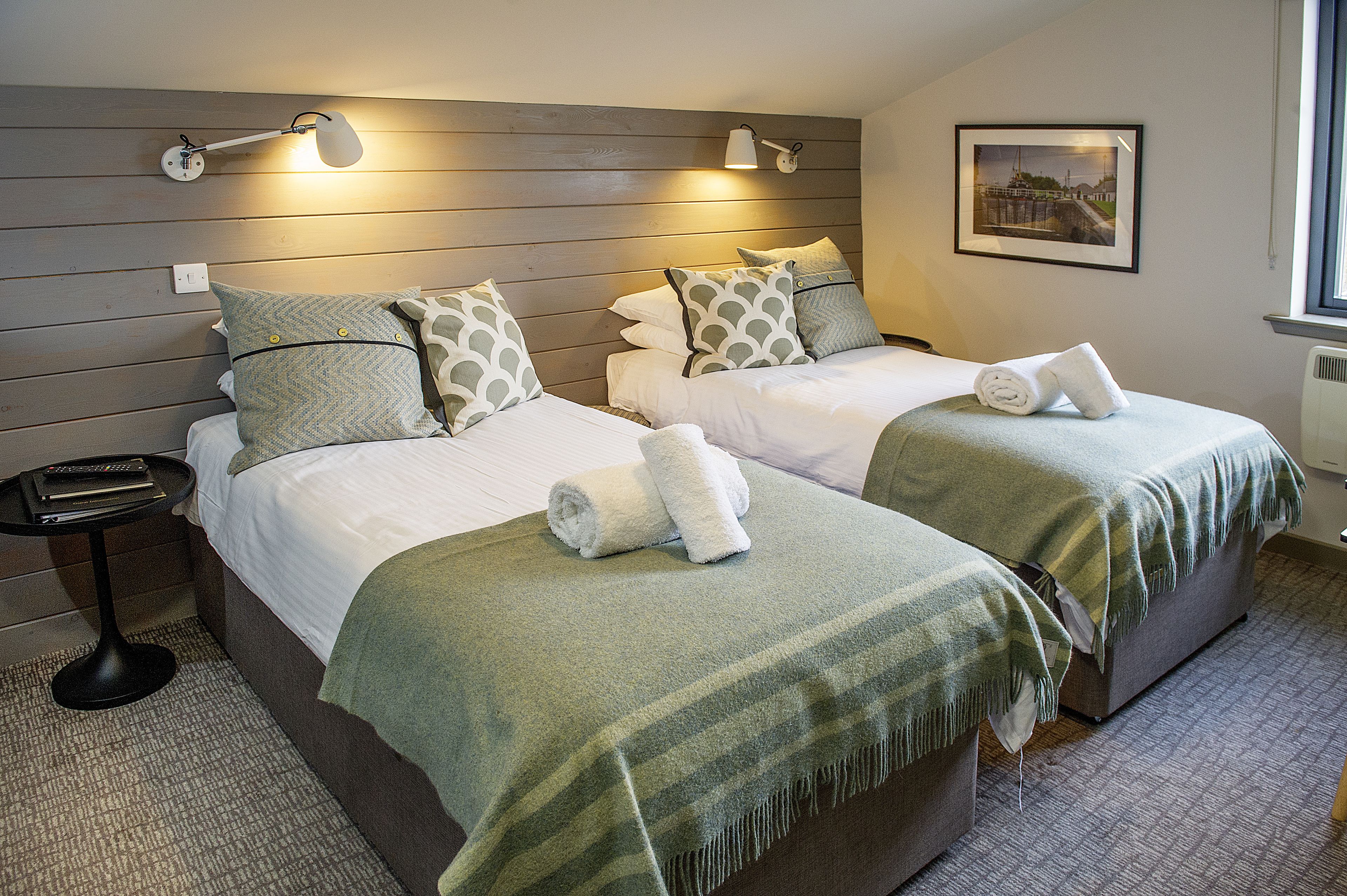 A hotel room with two beds, each with a green blanket and white bedding. There are wall-mounted lamps on either side of the beds, and a framed picture on the wall.