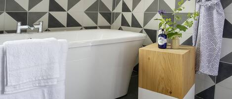 Ein modernes Badezimmer mit einer freistehenden Badewanne, einem Holztischchen mit Pflegeprodukten und einem waschmusterbehangten Duschbereich.