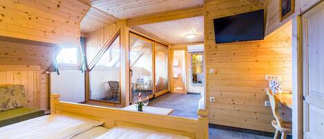 A wooden cabin room with a bed, a table with a wine glass, a chair, and a TV mounted on the wall.
