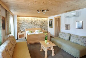 Una habitación acogedora con techo de madera, pared de piedra, cama con almohadas verdes, banco de madera, una mesita con un jarrón y un sofá verde.