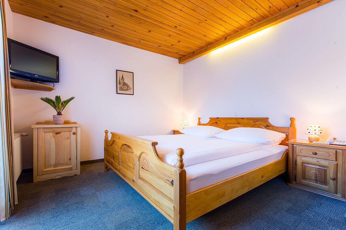 A wooden bed with a headboard, a bedside table with a lamp, a small cabinet, a television mounted on the wall, and a framed picture on the wall.