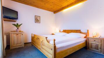 A wooden bed with a headboard, a bedside table with a lamp, a small cabinet, a television mounted on the wall, and a framed picture on the wall.