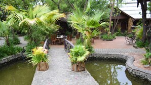 A garden with a stone bridge over a pond, surrounded by various plants and a wooden building with outdoor seating. - Bagan Central Hotel (Nyaung-U)