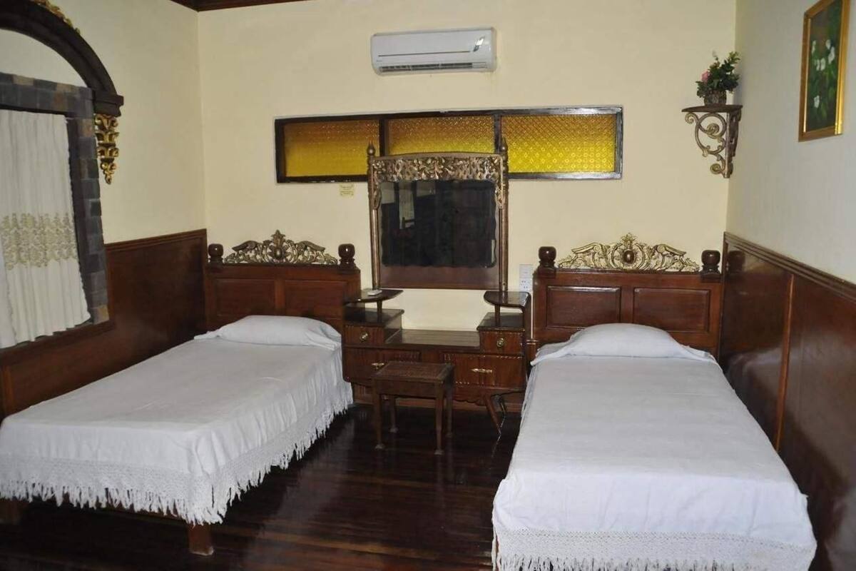 A room with two single beds, a wooden desk, a mirror, and an air conditioning unit.