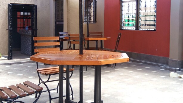 An outdoor patio area with wooden tables and benches, a large umbrella, and a red wall with windows.
