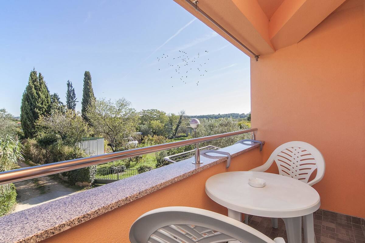 A balcony with a white table and chairs, a metal railing, and a view of trees and a path.