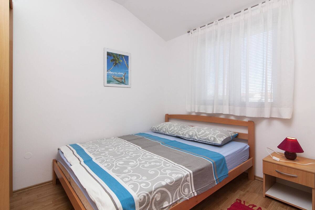 A single bed with a wooden frame, a bedside table with a lamp, and a window with white curtains.