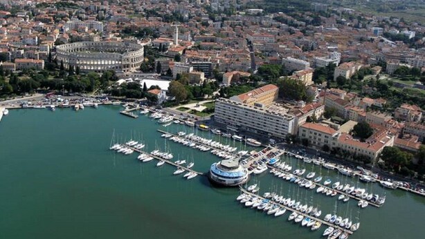 Vista aérea de una ciudad costera con un puerto deportivo lleno de barcos, rodeada de edificios históricos y una estructura circular destacada.
