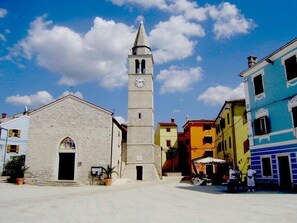 Ein Platz mit einer Kirche, einem Glockenturm, bunten Gebäuden und blauem Himmel.