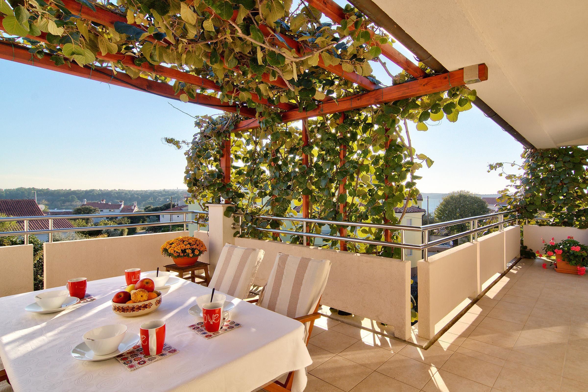 A terrace with a table set for two, featuring a fruit bowl, cups, and a flower vase.