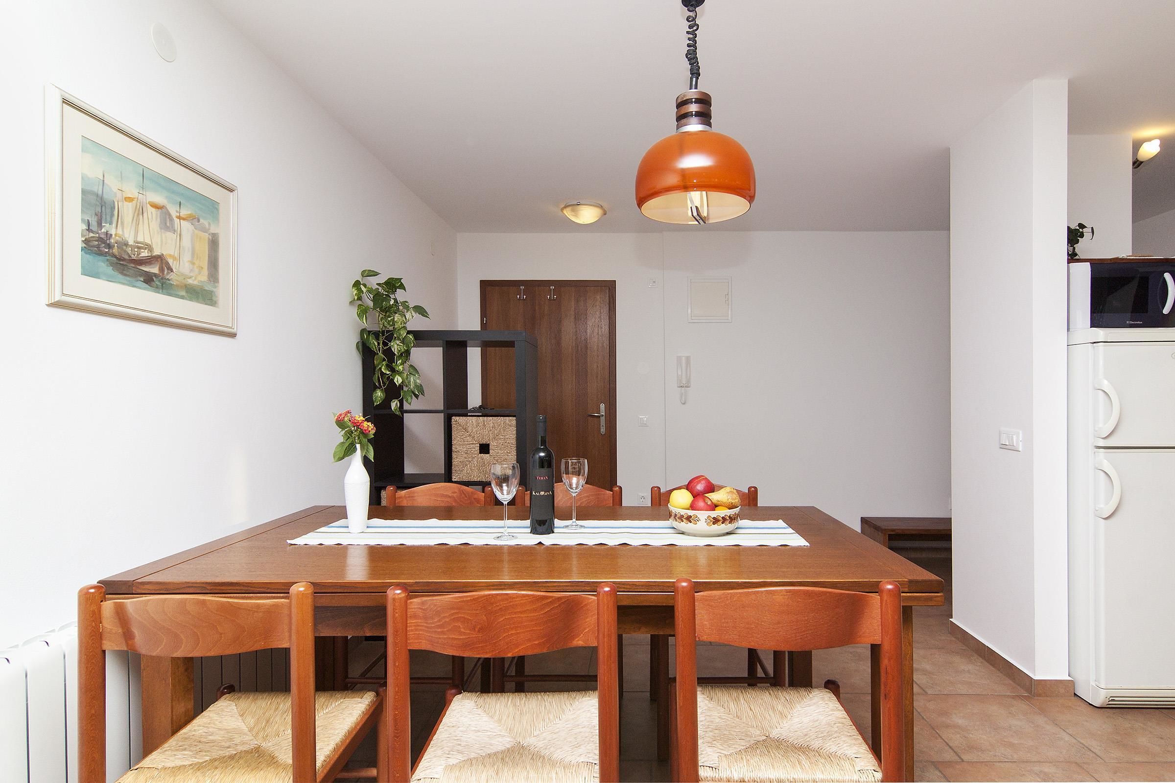 A dining area with a wooden table, chairs, a hanging light fixture, a framed painting, and a white refrigerator.