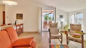 A spacious living room with a sofa, armchairs, and a dining area with a table and chairs. A balcony with a view of trees and buildings is visible through an open door. - Apartments Levan Ceja (Medulin)