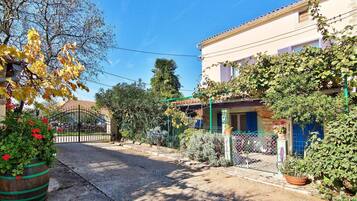 A quiet street with a house featuring a balcony, a garden with potted plants, and a clear blue sky.