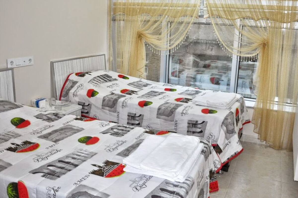 A hotel room with two beds covered in patterned bedding, a large window with curtains, and a radiator on the wall.