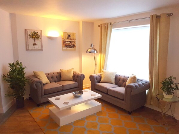A living room with two sofas, a coffee table, and a lamp. There are two potted plants and artwork on the walls. - Spacious Bright and Modern Living (Northampton)