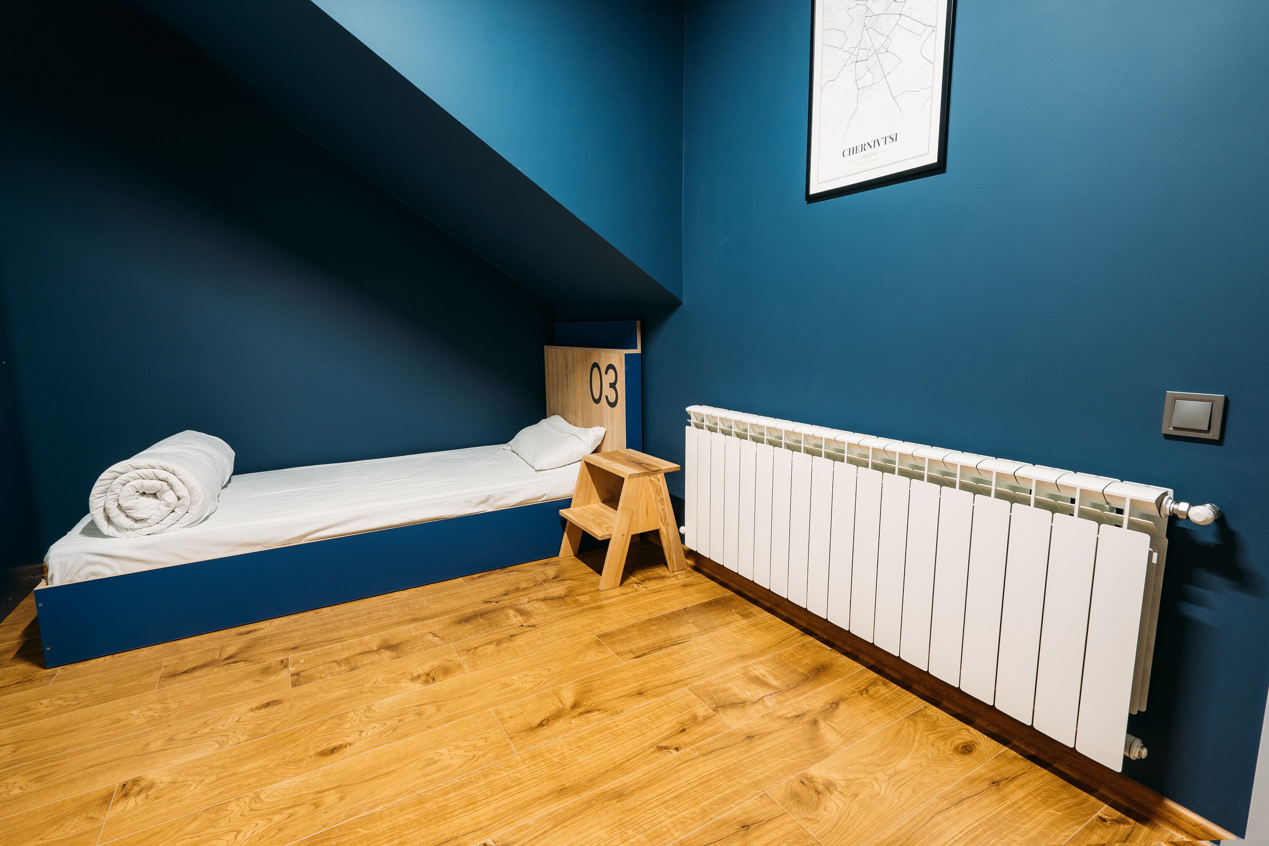 Um quarto pequeno com uma cama de solteiro, um banquinho de madeira e um radiador branco.