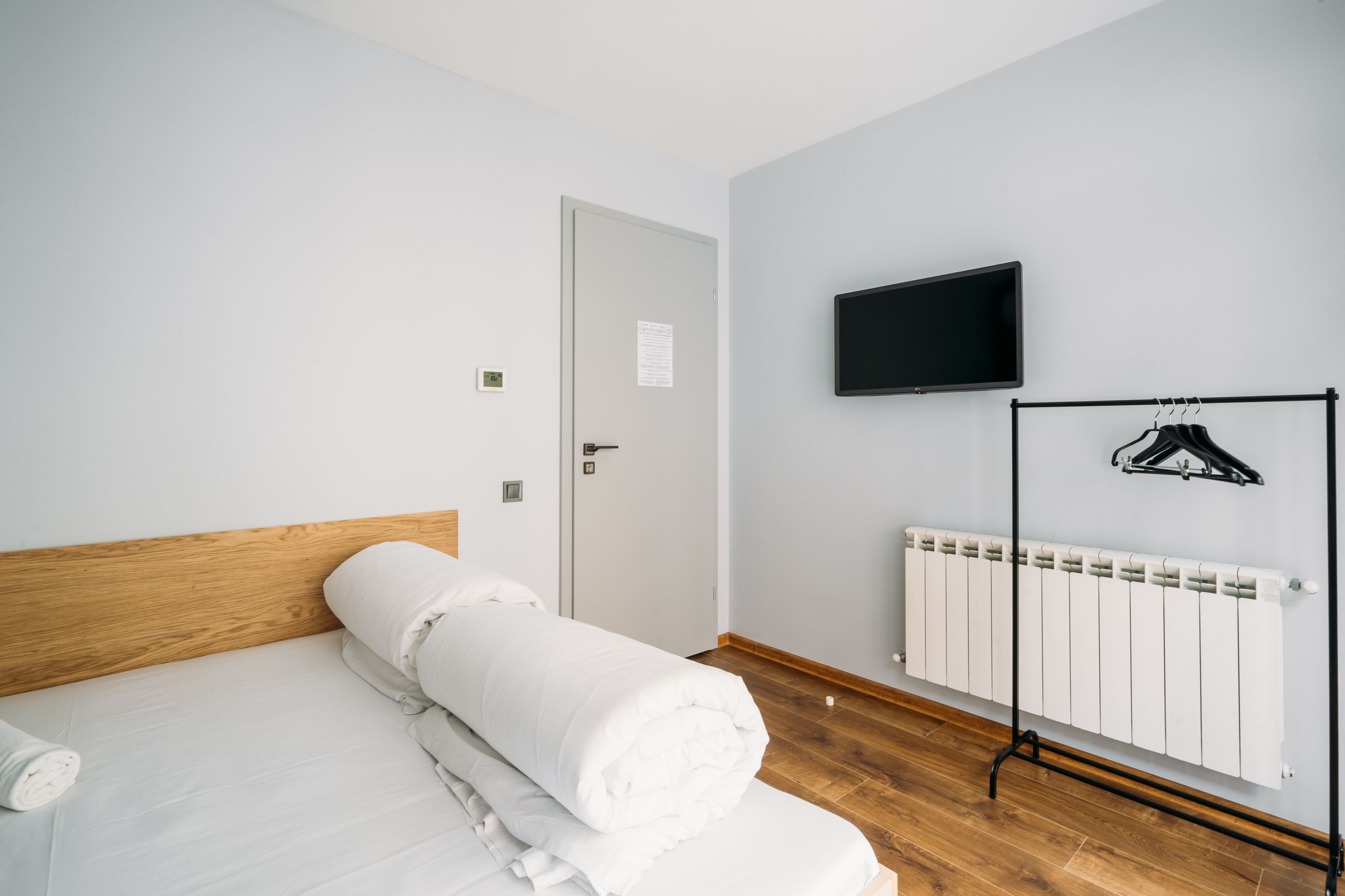 Une chambre d’hôtel avec une tête de lit en bois, un lit avec du linge de maison blanc, un téléviseur fixé au mur, un radiateur et un porte-vêtements.