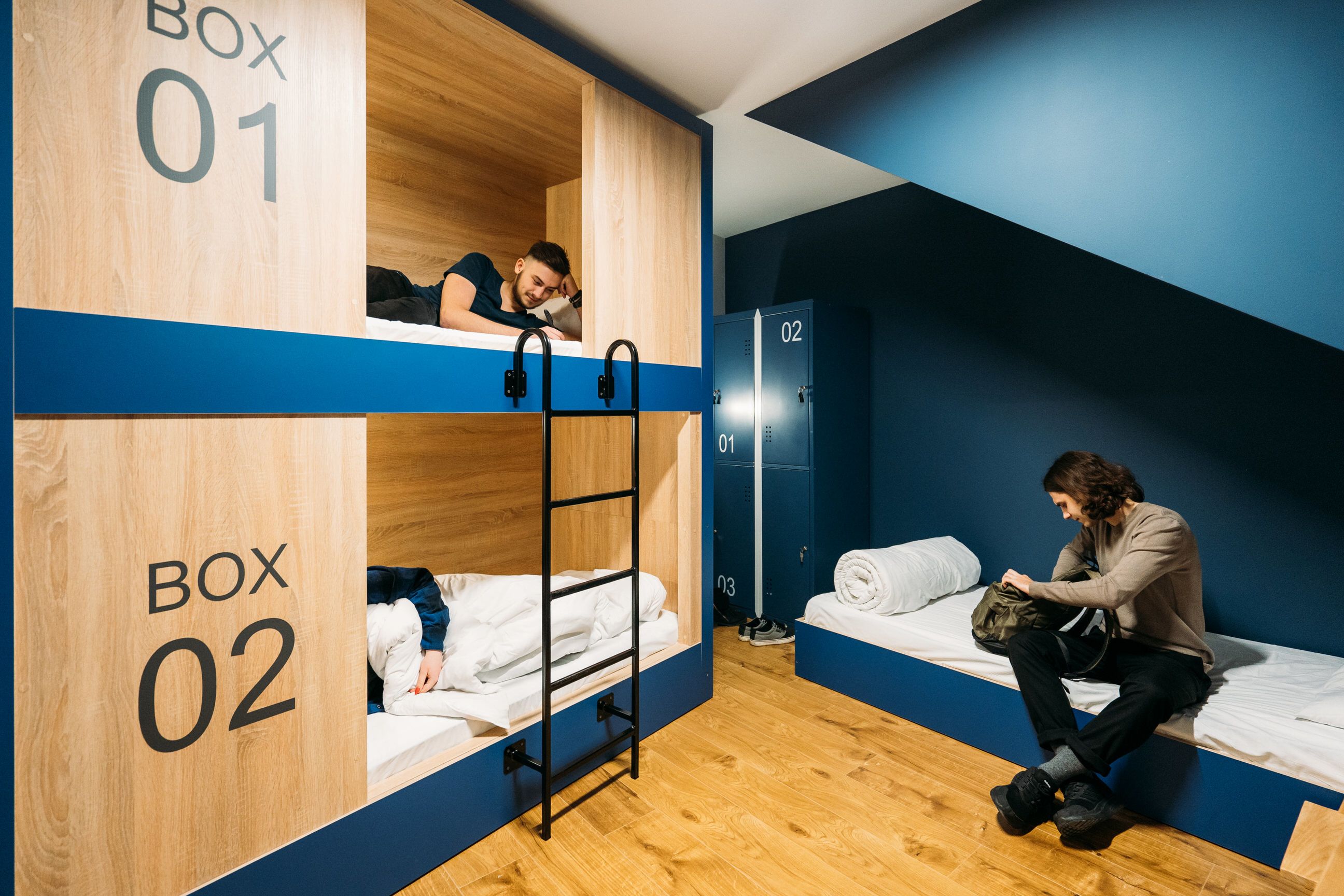 A dormitory room with bunk beds labeled Box 01 and Box 02, wooden floors, and a blue wall.