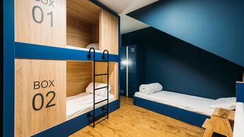 Ein Schlafsaalzimmer mit zwei Etagenbetten, beschriftet mit Box 01 und Box 02, Holzboden und einem kleinen Holzschemel.