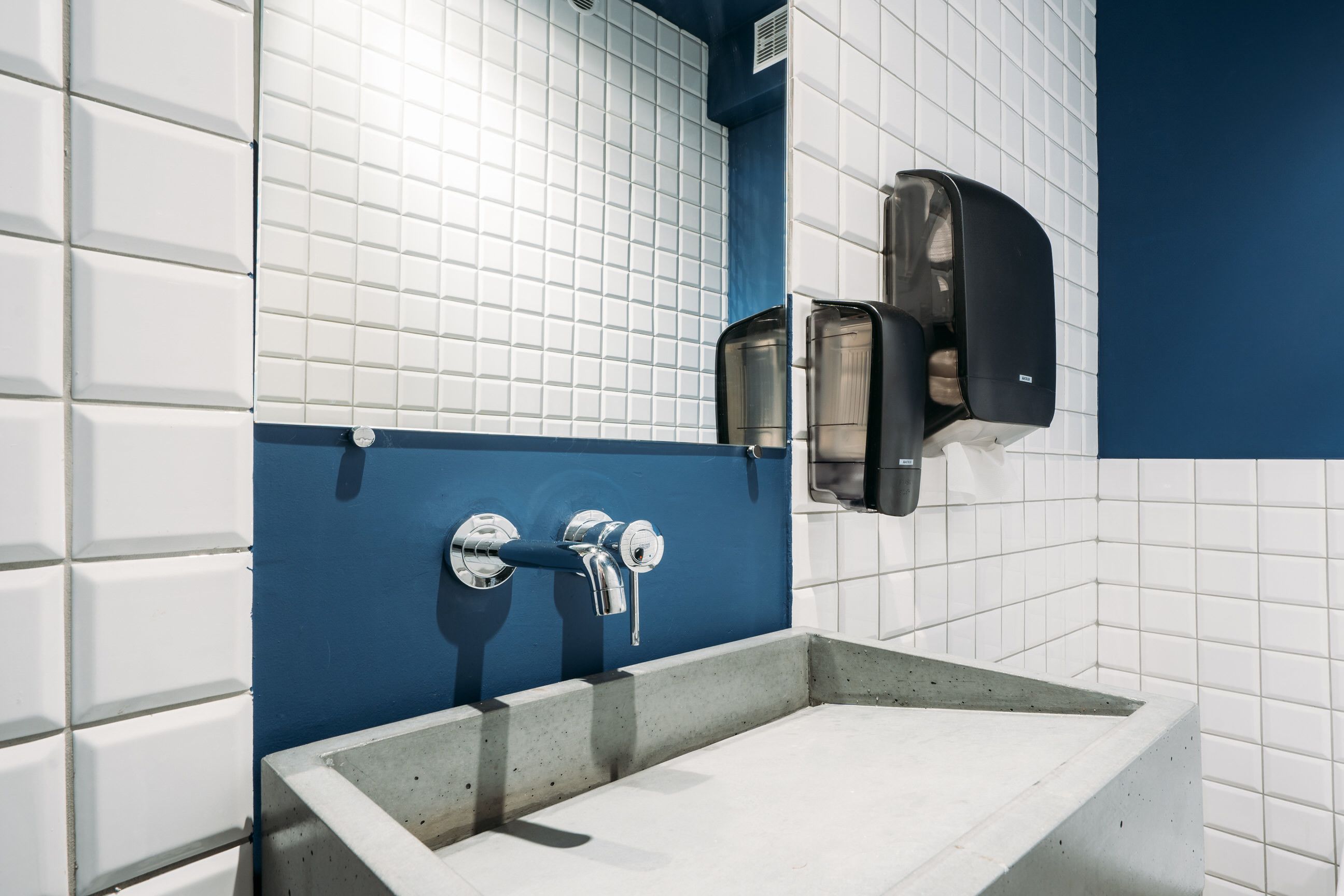 A bathroom with white tiled walls, a blue accent wall, and a concrete sink.