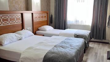 Een hotelkamer met drie bedden, houten hoofdeinden, een groot raam met gordijnen en een radiator.