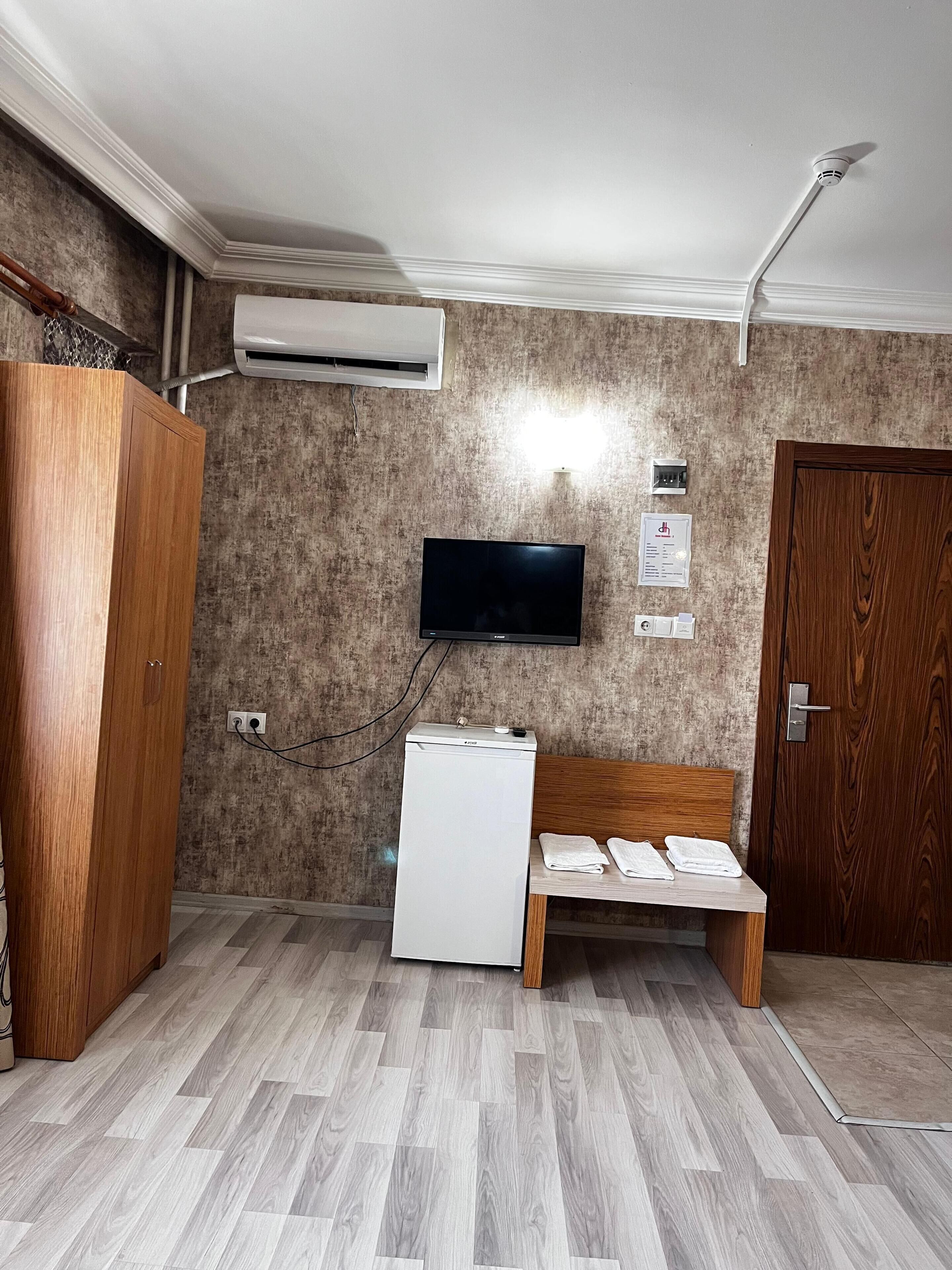 Una habitación con un armario de madera, un mini refrigerador blanco, un televisor de pantalla plana montado en la pared, un banco de madera y una puerta.