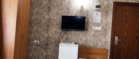 A room with a wooden wardrobe, a white mini-fridge, a flat-screen TV mounted on the wall, a wooden bench, and a door.