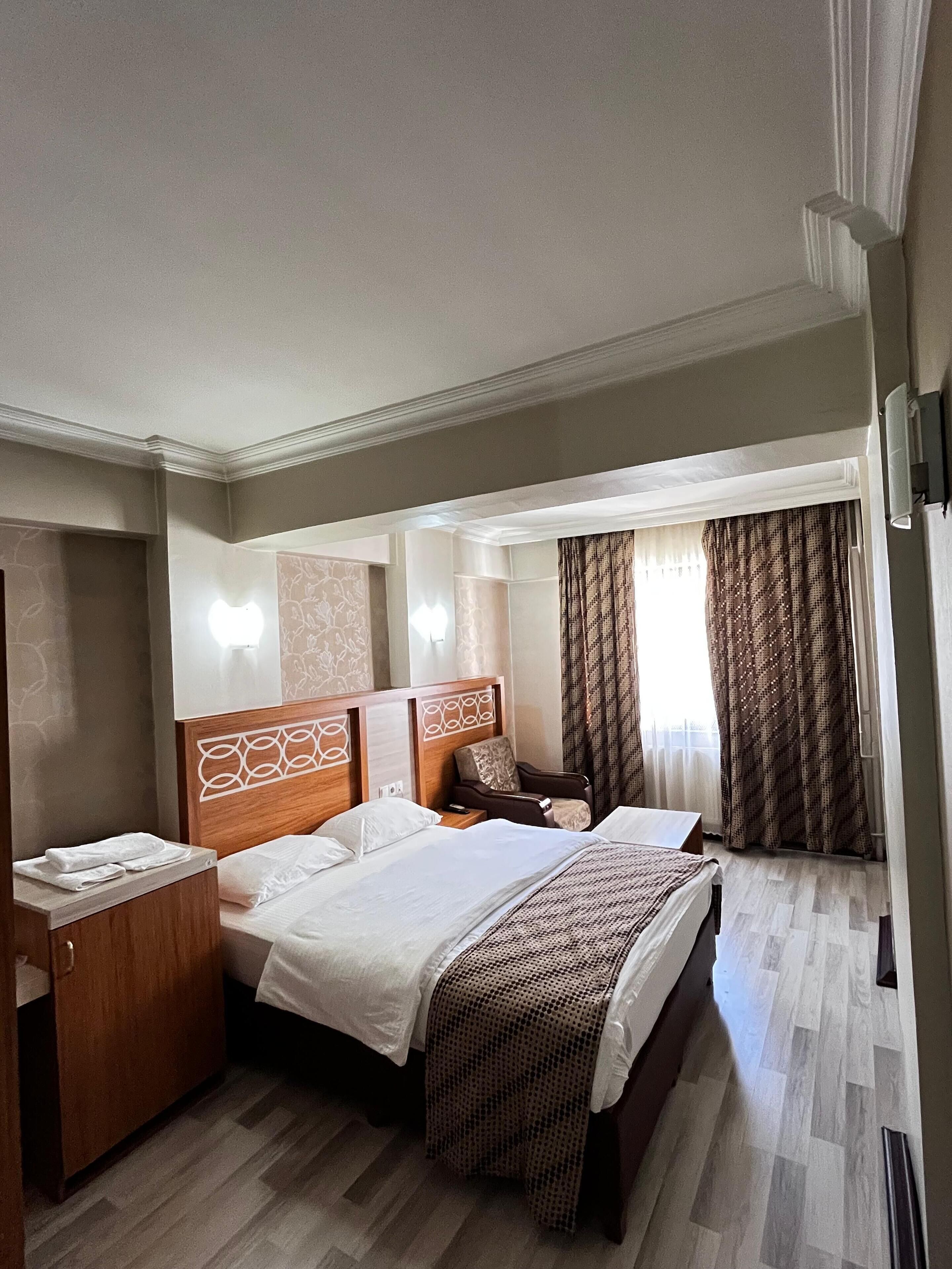 Una habitación de hotel con una cama grande, cabecera de madera, mesita de noche, una silla y una ventana con cortinas.