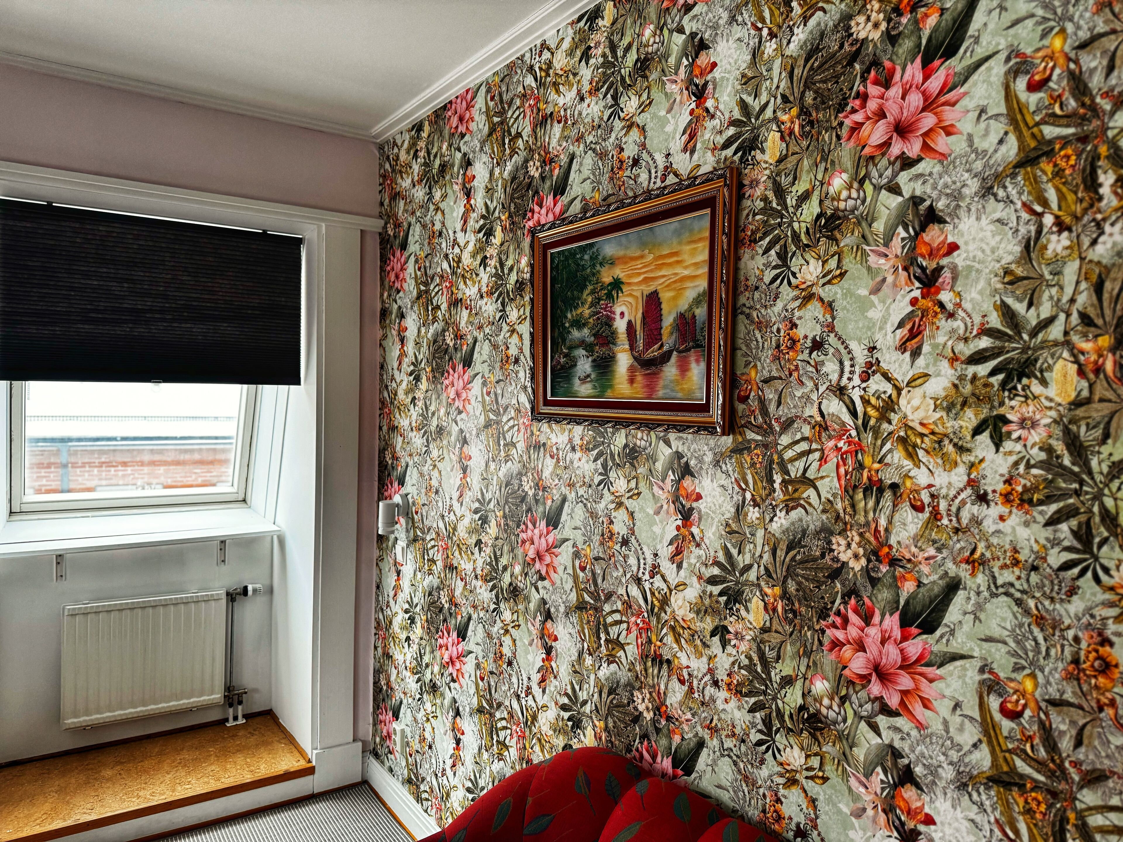 A room with floral wallpaper, a framed picture, a window with a view, and a radiator.