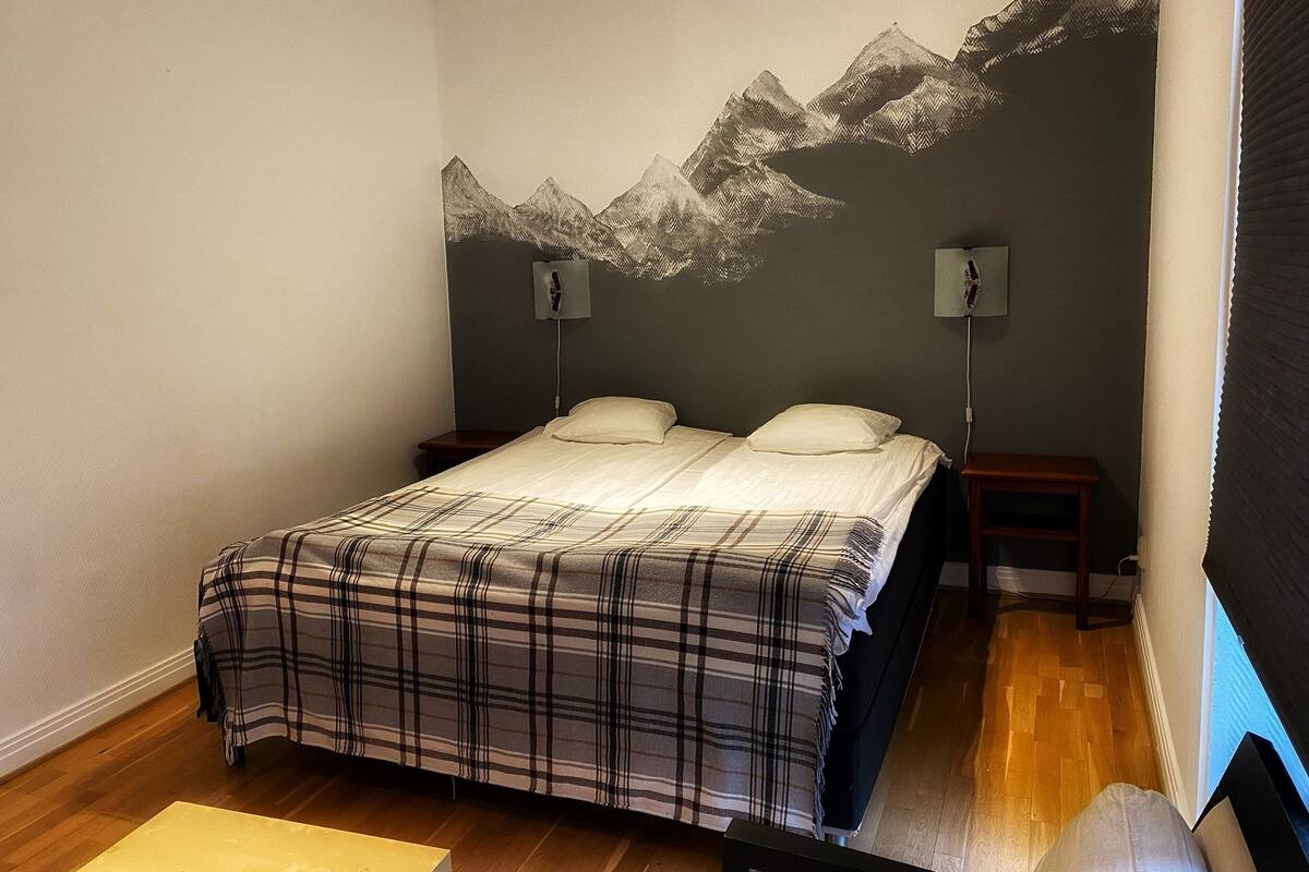 A bedroom with a bed, two bedside tables, a wall-mounted lamp, and a wooden floor.