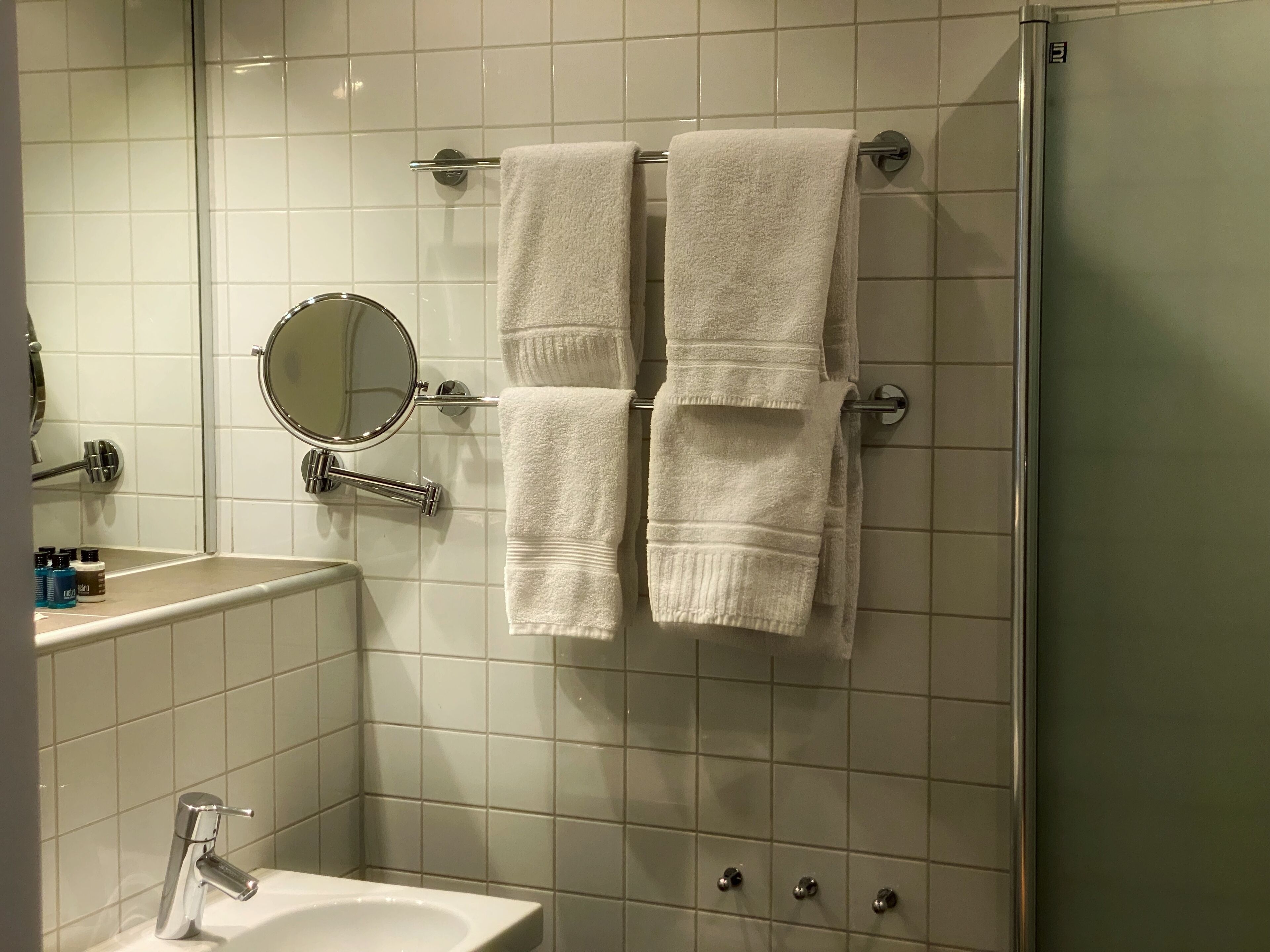 Un baño con azulejos blancos, un lavabo, un espejo y dos toallas colgadas en un tendedero.