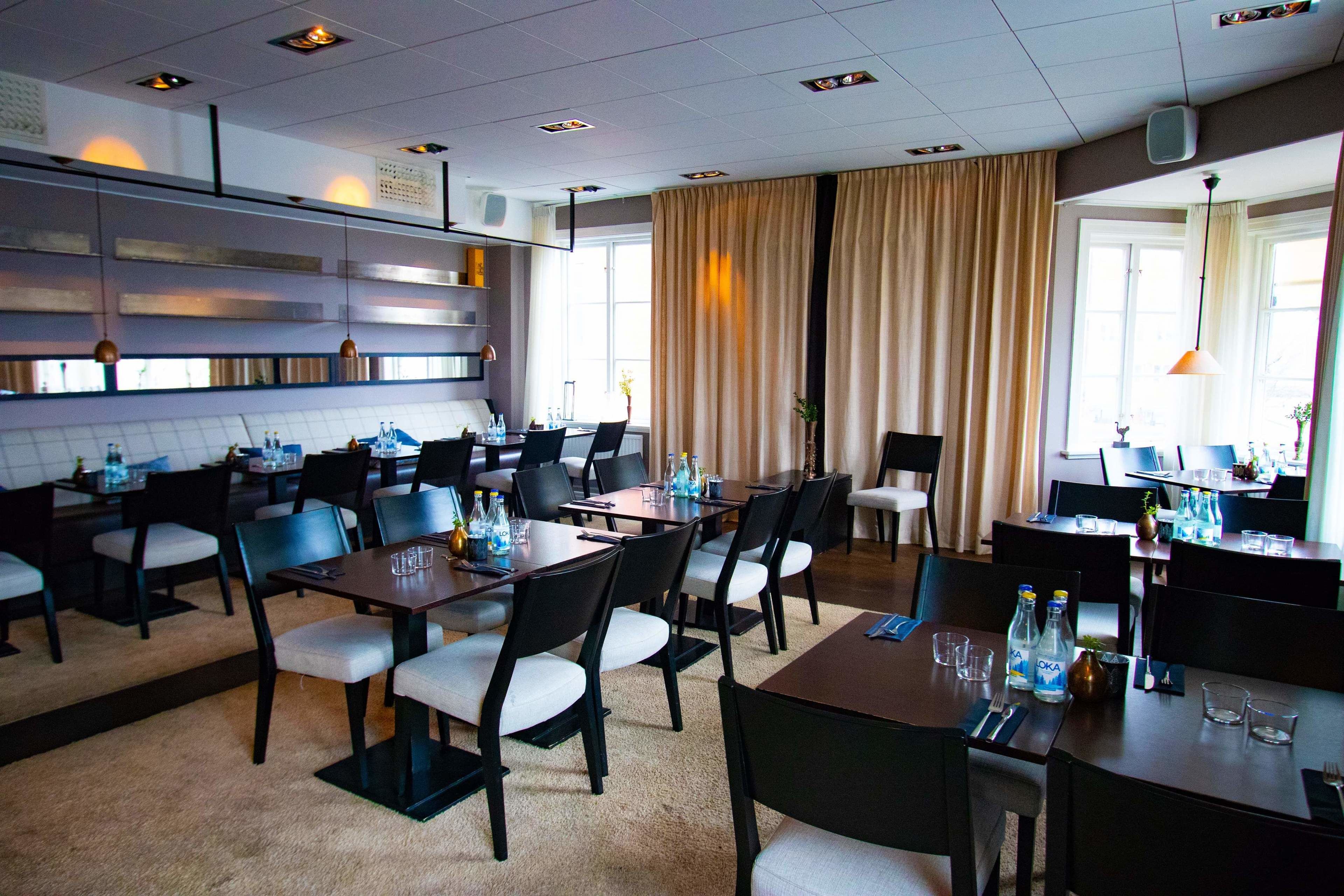 En hotellrestaurang med flera bord och stolar, ett köksområde och stora fönster med gardiner.