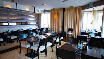 En hotellrestaurang med flera bord och stolar, ett köksområde och stora fönster med gardiner.
