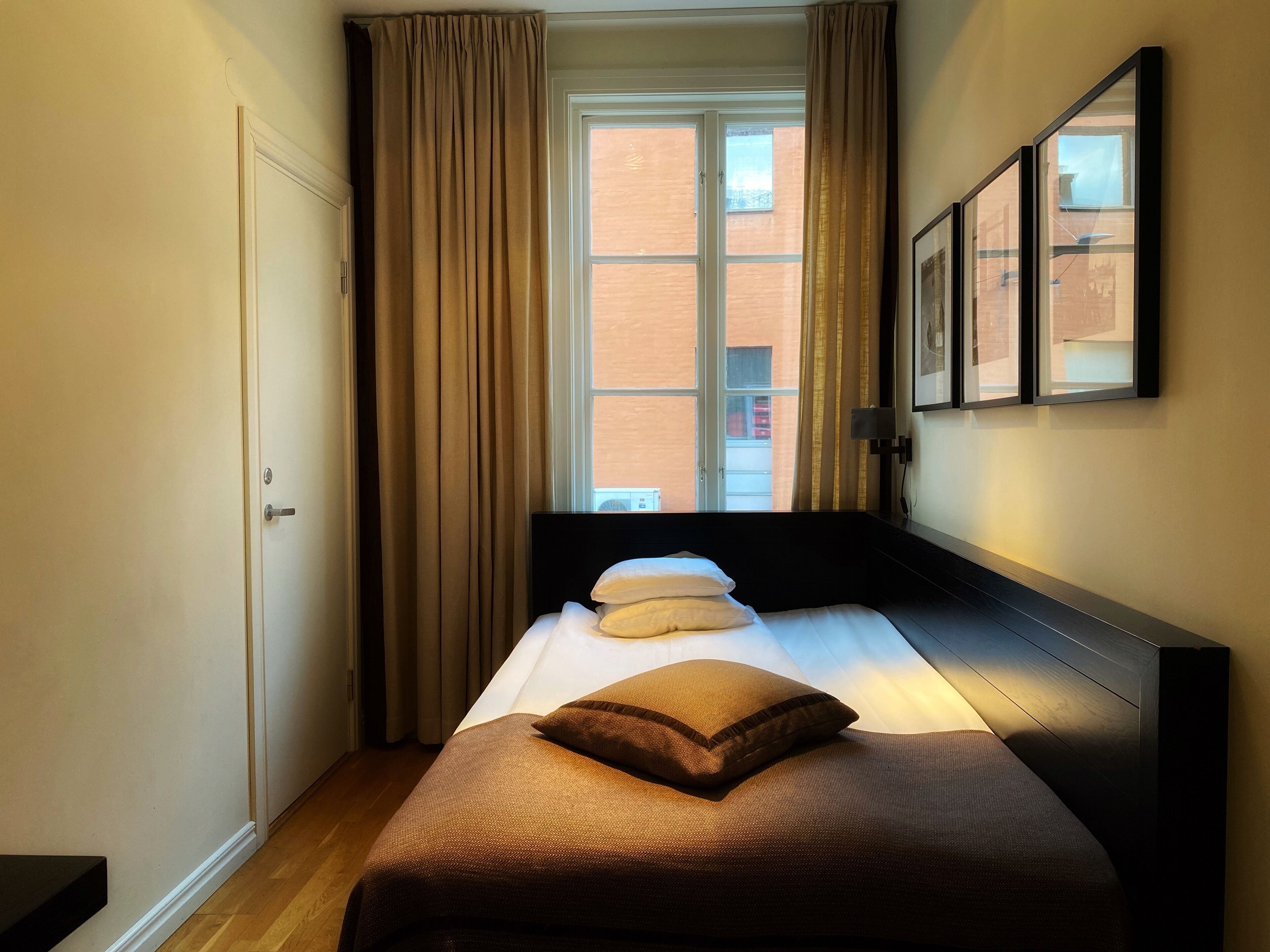 Quarto de hotel com cama, janela com cortinas, porta e um quadro na parede.