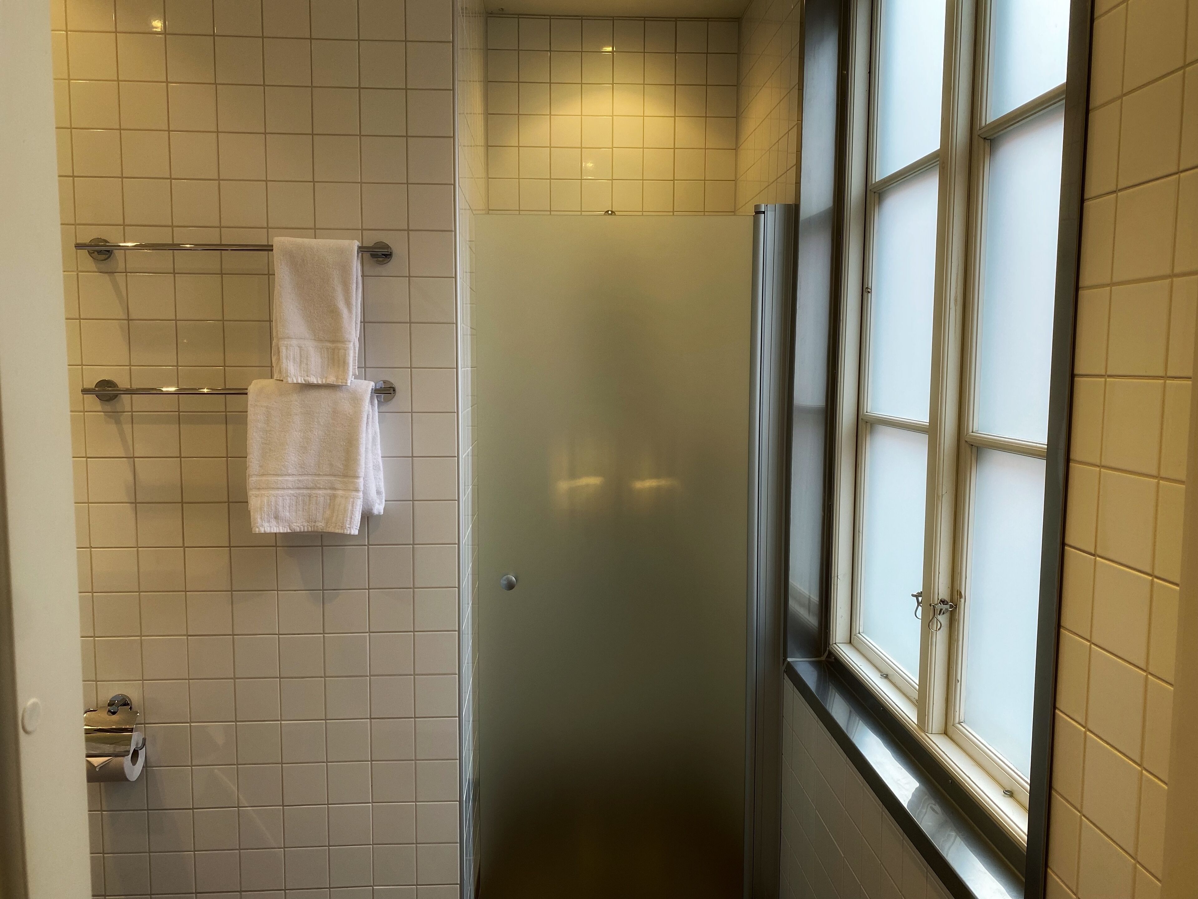 A bathroom with a tiled wall, a towel rack with two towels, a toilet paper holder, and a window with frosted glass.