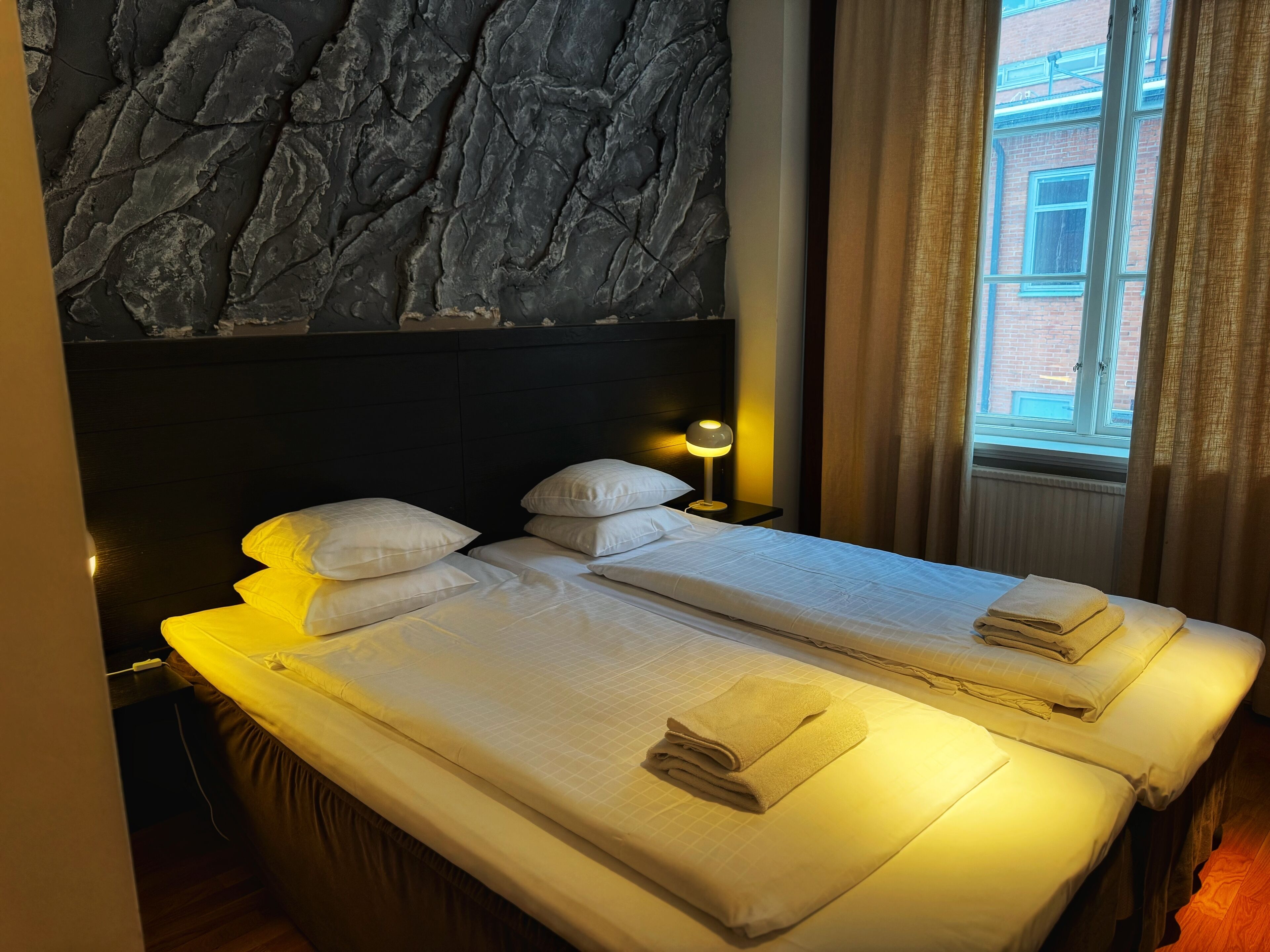 Ein Doppelbett mit weißen Laken und zwei Handtüchern, ein Nachttisch mit Lampe sowie eine Steinwand.