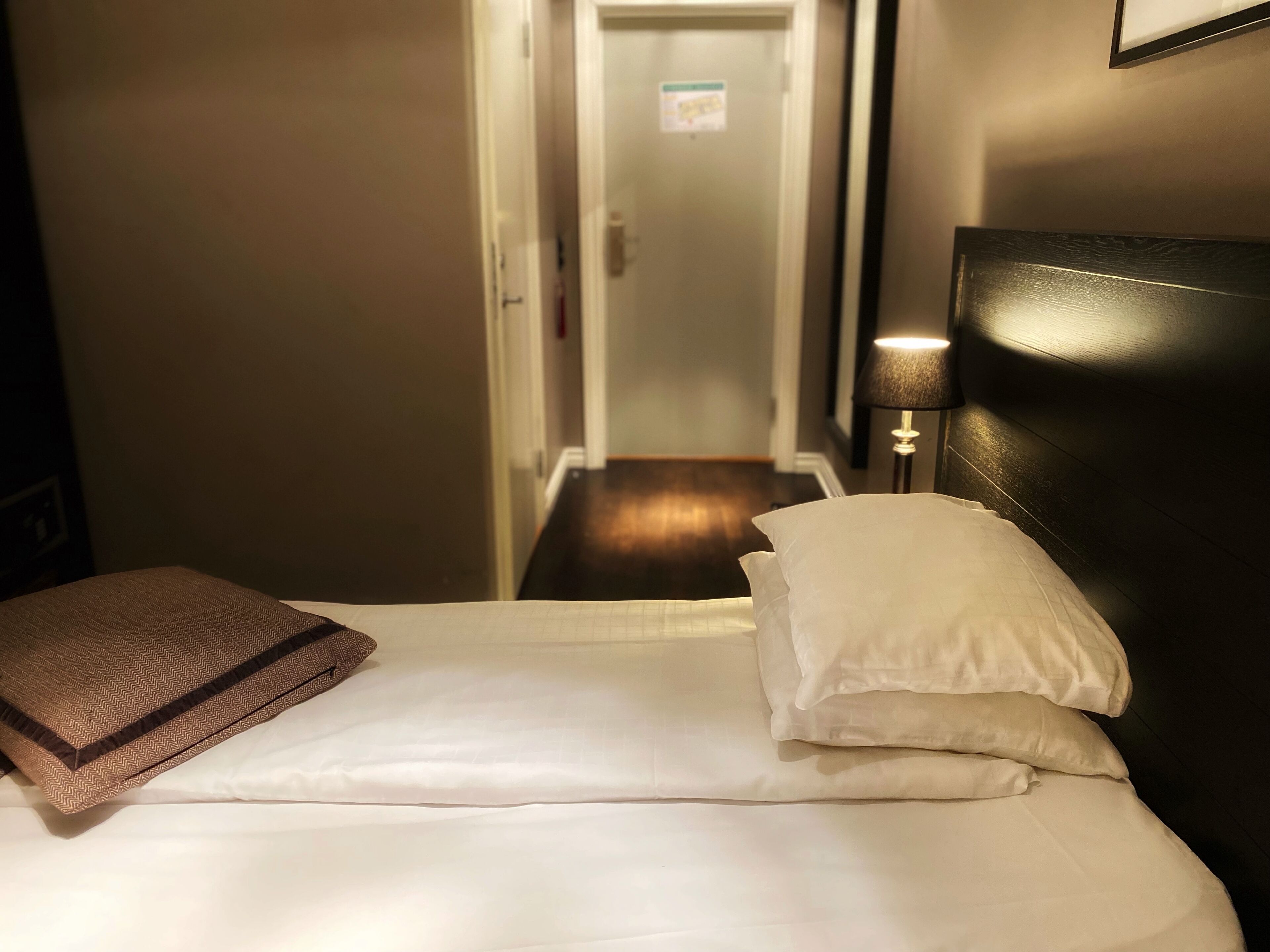 A hotel room with a bed, pillows, a lamp, and a door leading to a hallway.