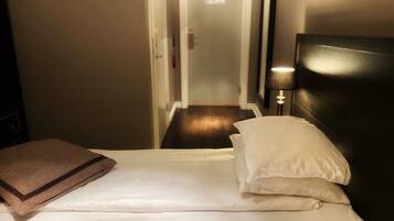 A hotel room with a bed, pillows, a lamp, and a door leading to a hallway.