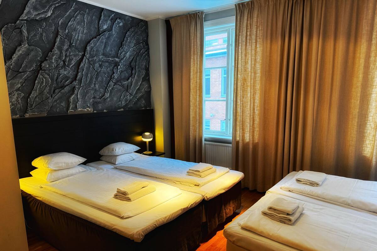 A hotel room with two beds, a stone wall, a window with curtains, and a nightstand with a lamp.