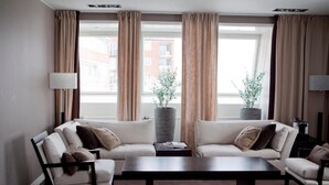 Un salon moderne avec deux canapés, une table basse et une lampe. De grandes fenêtres sont ornées de rideaux, et deux plantes en pot sont posées sur le rebord de la fenêtre.