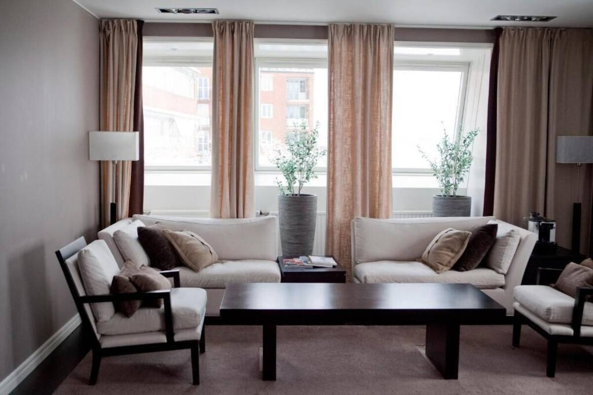 A modern living room with two sofas, a coffee table, and a lamp. There are large windows with curtains, and two potted plants on the windowsill.