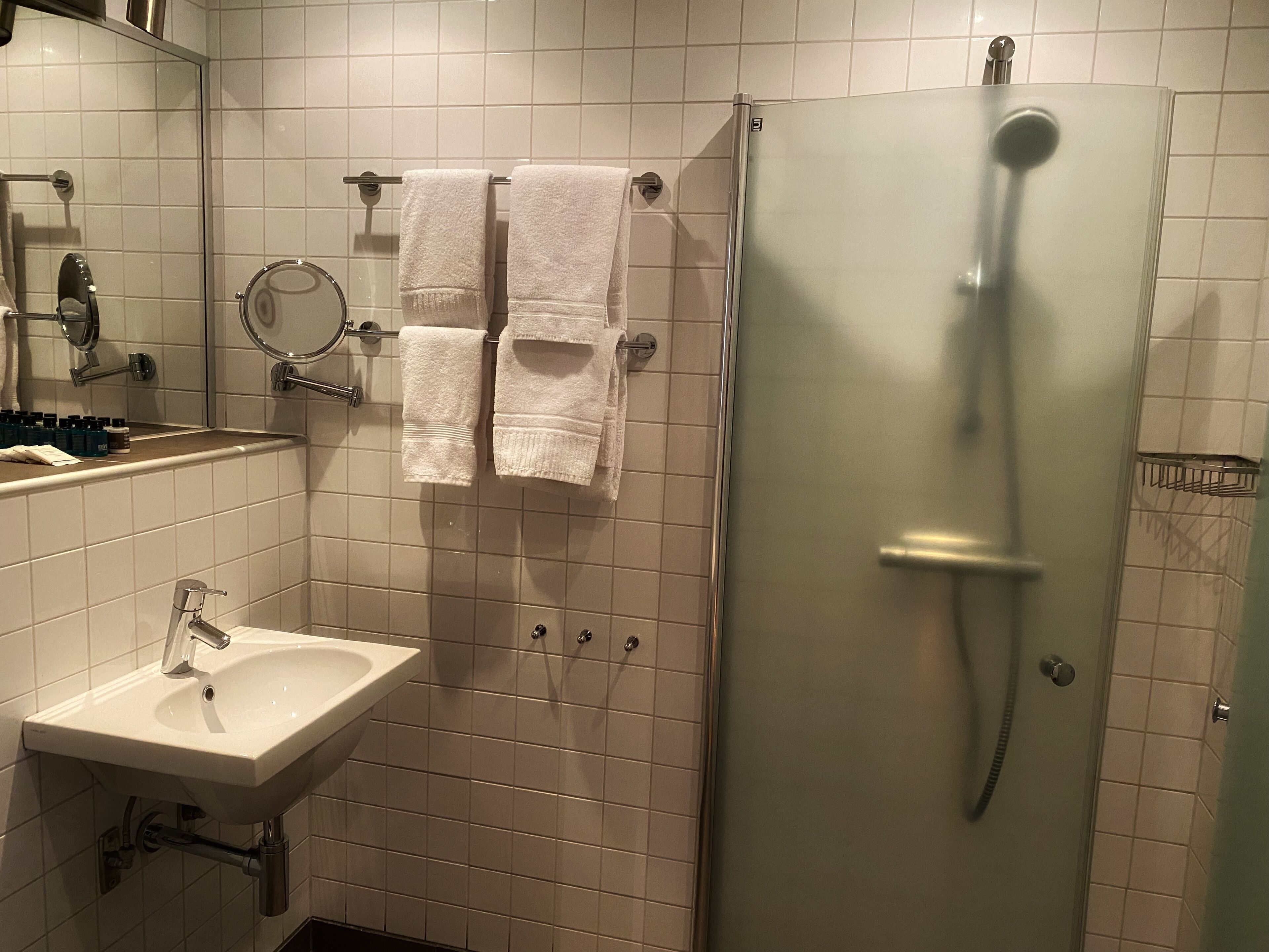 Un baño con lavamanos, dos toallas colgadas en un perchero, un espejo y una cabina de ducha.